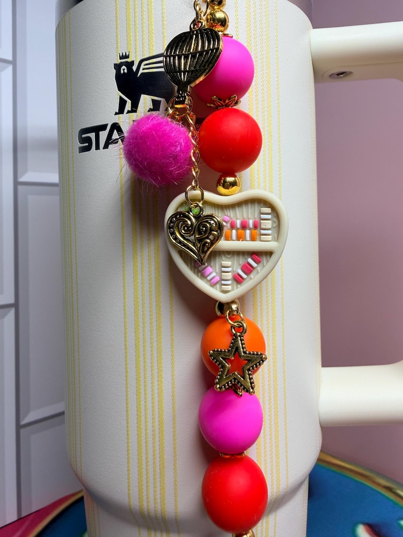 K&ouml;nnte beinhalten: Ein dekorativer Charm mit einer goldenen Kette, einem rosa Pompon und bunten Perlen. Der Charm hat einen herzf&ouml;rmigen Anh&auml;nger mit einem B&uuml;cherregal-Design und einem goldenen Stern. Der Charm ist an einem wei&szlig;en und beigen Stanley-Becher befestigt.