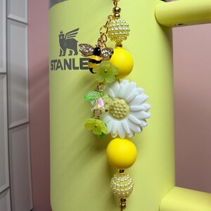 May include: A decorative keychain featuring a gold-tone metal clasp, yellow and white beads, a bee charm with clear crystals, and a white daisy flower charm. The keychain is attached to a yellow tumbler with the word STANLEY printed in black.