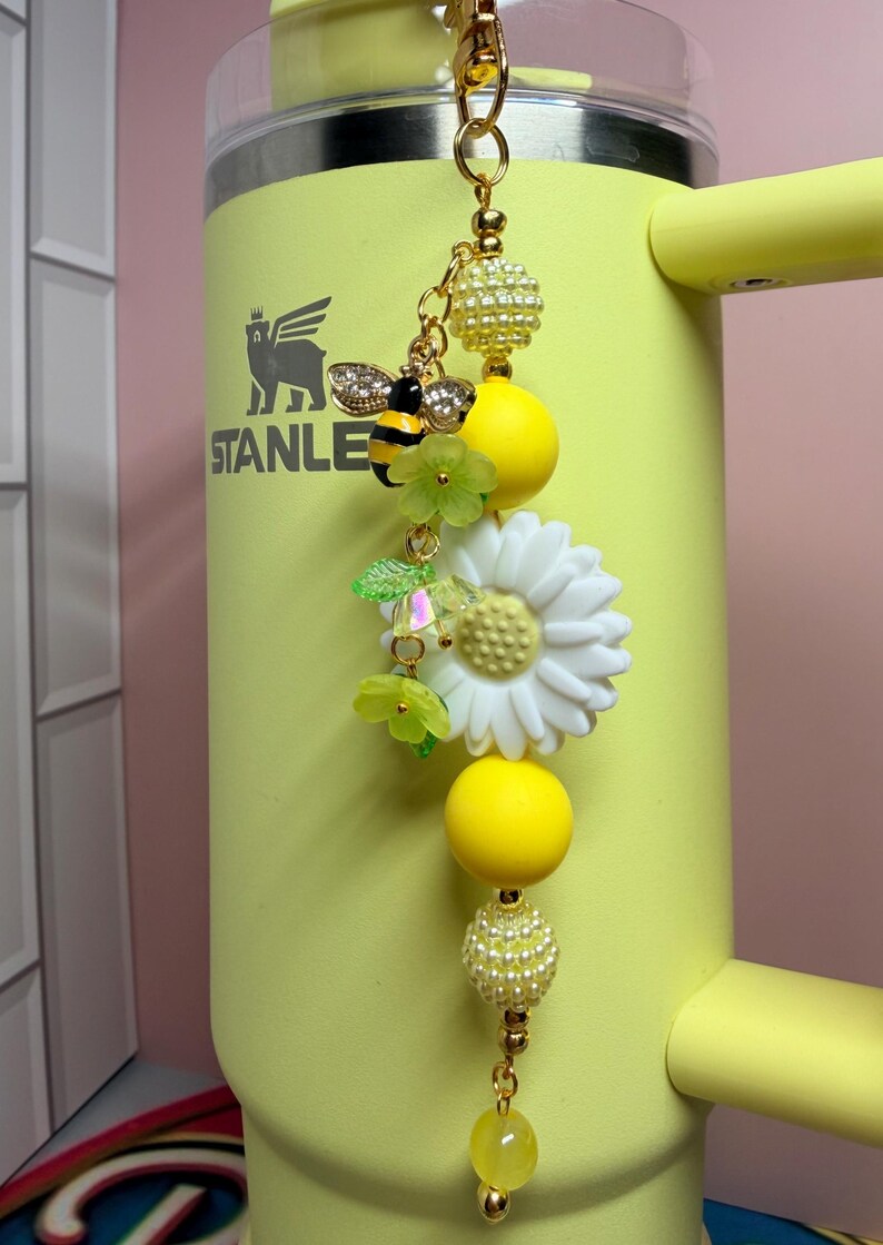 May include: A decorative keychain featuring a gold-tone metal clasp, yellow and white beads, a bee charm with clear crystals, and a white daisy flower charm. The keychain is attached to a yellow tumbler with the word STANLEY printed in black.