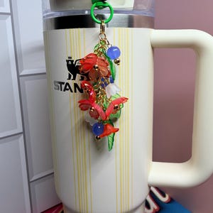 May include: A cream-colored insulated tumbler with a green clip-on keychain. The keychain features a cluster of red, white, and blue flower charms with gold accents. The tumbler has a white handle and the word "STAN" printed on the side.
