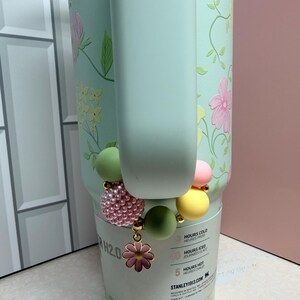 May include: A light green Stanley H2.0 water bottle with a floral design and a beaded charm bracelet. The bracelet features pink, yellow, and green beads, and a pink flower charm. The bottle has a handle.