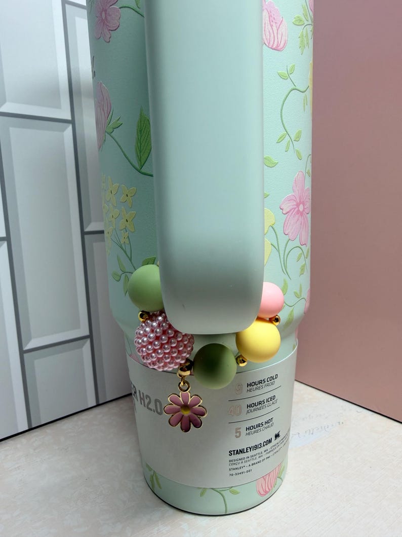 May include: A light green Stanley H2.0 water bottle with a floral design and a beaded charm bracelet. The bracelet features pink, yellow, and green beads, and a pink flower charm. The bottle has a handle.