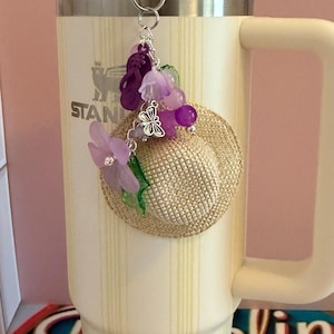May include: A decorative charm featuring a miniature straw hat, purple flower, and beads, attached to a silver clip. The charm is hanging on a cream-coloured insulated tumbler with the word "STANLEY" visible.