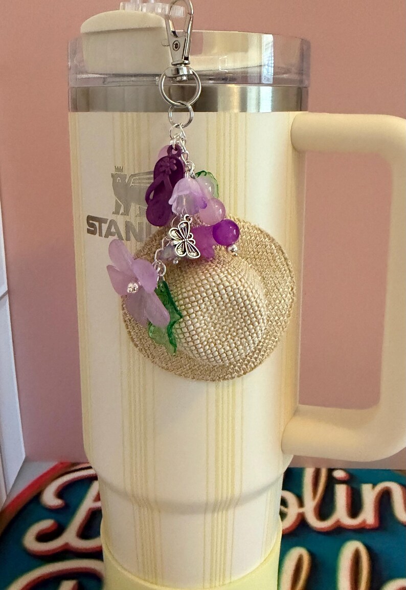 May include: A decorative charm featuring a miniature straw hat, purple flower, and beads, attached to a silver clip. The charm is hanging on a cream-coloured insulated tumbler with the word "STANLEY" visible.