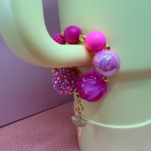 Op de afbeelding: Een armband met grote, ronde roze en paarse kralen, afgewisseld met goudkleurige afstandhouders. Het heeft een bedel in de vorm van een ballerina. E&eacute;n kraal is bedekt met roze strass-steentjes. De armband wordt gepresenteerd op een lichtgroen oppervlak.