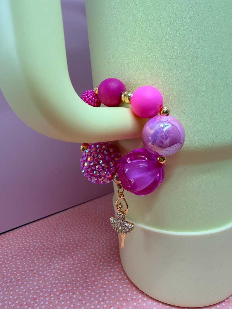 Op de afbeelding: Een armband met grote, ronde roze en paarse kralen, afgewisseld met goudkleurige afstandhouders. Het heeft een bedel in de vorm van een ballerina. E&eacute;n kraal is bedekt met roze strass-steentjes. De armband wordt gepresenteerd op een lichtgroen oppervlak.