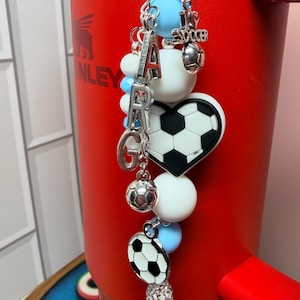 Puede incluir: Un dije decorativo para vaso con cuentas de silicona azul claro y blancas, letras plateadas que forman ARG, un dije de coraz&oacute;n con un bal&oacute;n de f&uacute;tbol, un dije de bal&oacute;n peque&ntilde;o y un colgante redondo de bal&oacute;n en una cadena plateada.