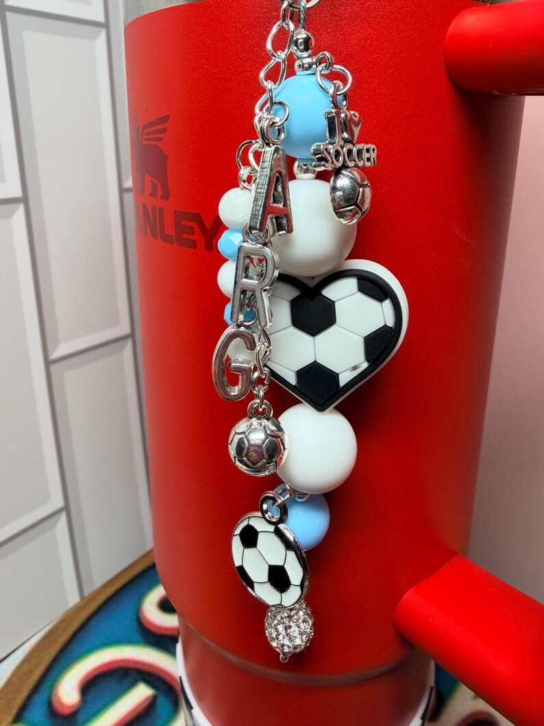 Puede incluir: Un dije decorativo para vaso con cuentas de silicona azul claro y blancas, letras plateadas que forman ARG, un dije de coraz&oacute;n con un bal&oacute;n de f&uacute;tbol, un dije de bal&oacute;n peque&ntilde;o y un colgante redondo de bal&oacute;n en una cadena plateada.
