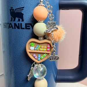 K&ouml;nnte beinhalten: Ein Schl&uuml;sselanh&auml;nger mit Perlen mit einem herzf&ouml;rmigen B&uuml;cherregal-Anh&auml;nger, einem Kronen-Anh&auml;nger und einem kleinen orangefarbenen Pompon. Der Schl&uuml;sselanh&auml;nger hat orangefarbene, cremefarbene und transparente Perlen. Der Schl&uuml;sselanh&auml;nger ist an einer blauen Stanley-Tasse befestigt.