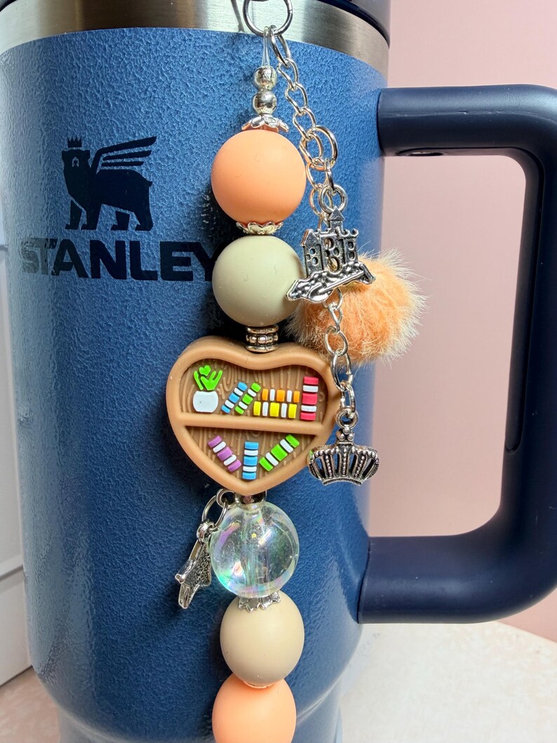 K&ouml;nnte beinhalten: Ein Schl&uuml;sselanh&auml;nger mit Perlen mit einem herzf&ouml;rmigen B&uuml;cherregal-Anh&auml;nger, einem Kronen-Anh&auml;nger und einem kleinen orangefarbenen Pompon. Der Schl&uuml;sselanh&auml;nger hat orangefarbene, cremefarbene und transparente Perlen. Der Schl&uuml;sselanh&auml;nger ist an einer blauen Stanley-Tasse befestigt.