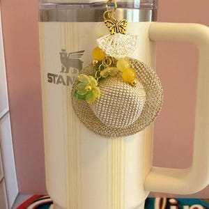 May include: A cream-coloured Stanley mug with a gold charm. The charm features a miniature straw hat, yellow beads, and a butterfly pendant. The mug has a handle and a lid with a straw. The mug has the word "STANLEY" printed on it.