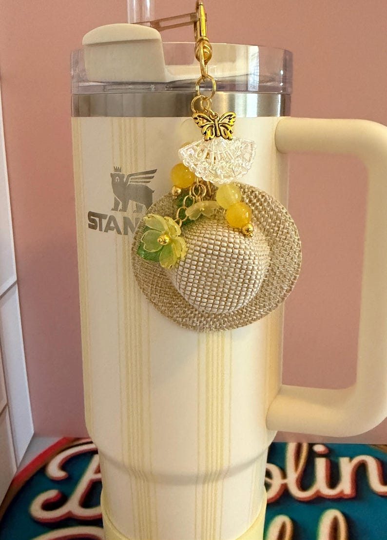 May include: A cream-coloured Stanley mug with a gold charm. The charm features a miniature straw hat, yellow beads, and a butterfly pendant. The mug has a handle and a lid with a straw. The mug has the word "STANLEY" printed on it.