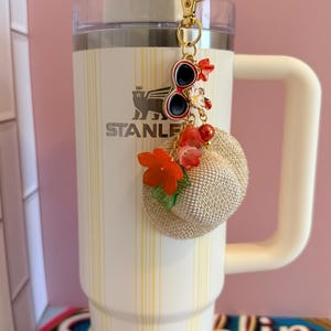 May include: A white Stanley tumbler with a yellow stripe design and a matching handle. A gold keychain with sunglasses and a small straw hat charm hangs from the tumbler. The tumbler has the word "STANLEY" printed on it.