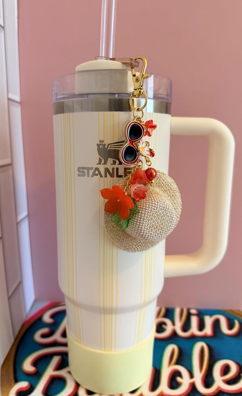 May include: A white Stanley tumbler with a yellow stripe design and a matching handle. A gold keychain with sunglasses and a small straw hat charm hangs from the tumbler. The tumbler has the word "STANLEY" printed on it.