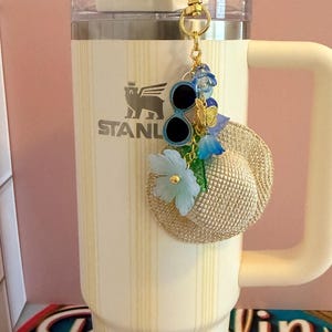 May include: A cream Stanley tumbler with a straw and handle, decorated with a gold charm including sunglasses, a miniature straw hat, blue flowers, and a butterfly. The tumbler has the "STANLEY" logo.