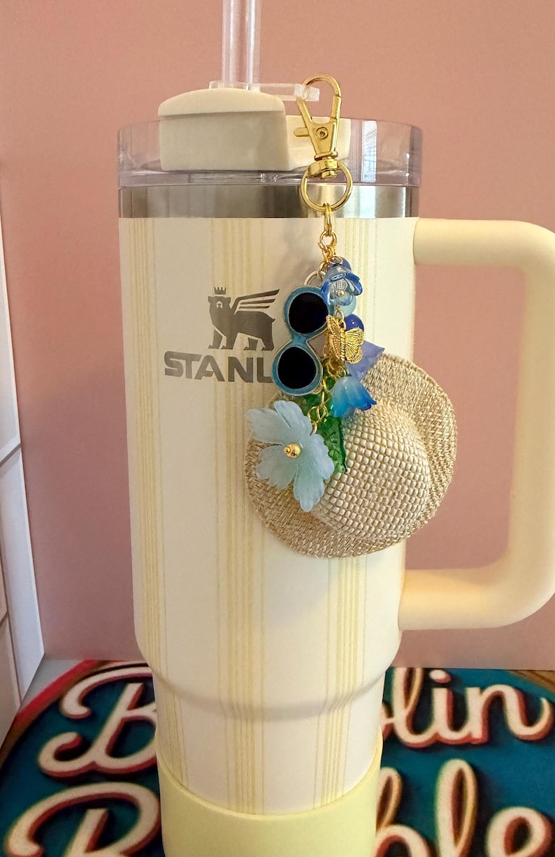 May include: A cream Stanley tumbler with a straw and handle, decorated with a gold charm including sunglasses, a miniature straw hat, blue flowers, and a butterfly. The tumbler has the "STANLEY" logo.
