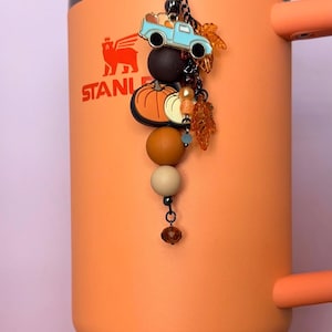 Puede incluir: Un vaso naranja mate con el logotipo de Stanley y un dije colgante que presenta un camión azul claro, dijes de calabazas naranjas y blancas, dijes de hojas de color ámbar translúcido y una serie de cuentas de silicona redondas en tonos marrón, beige y naranja.