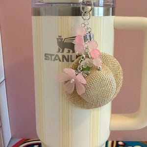 May include: A cream-coloured Stanley tumbler with a straw and a decorative keychain. The keychain features a small straw hat, pink flower accents, and pink bead embellishments. The tumbler has the Stanley logo and a yellow striped pattern.