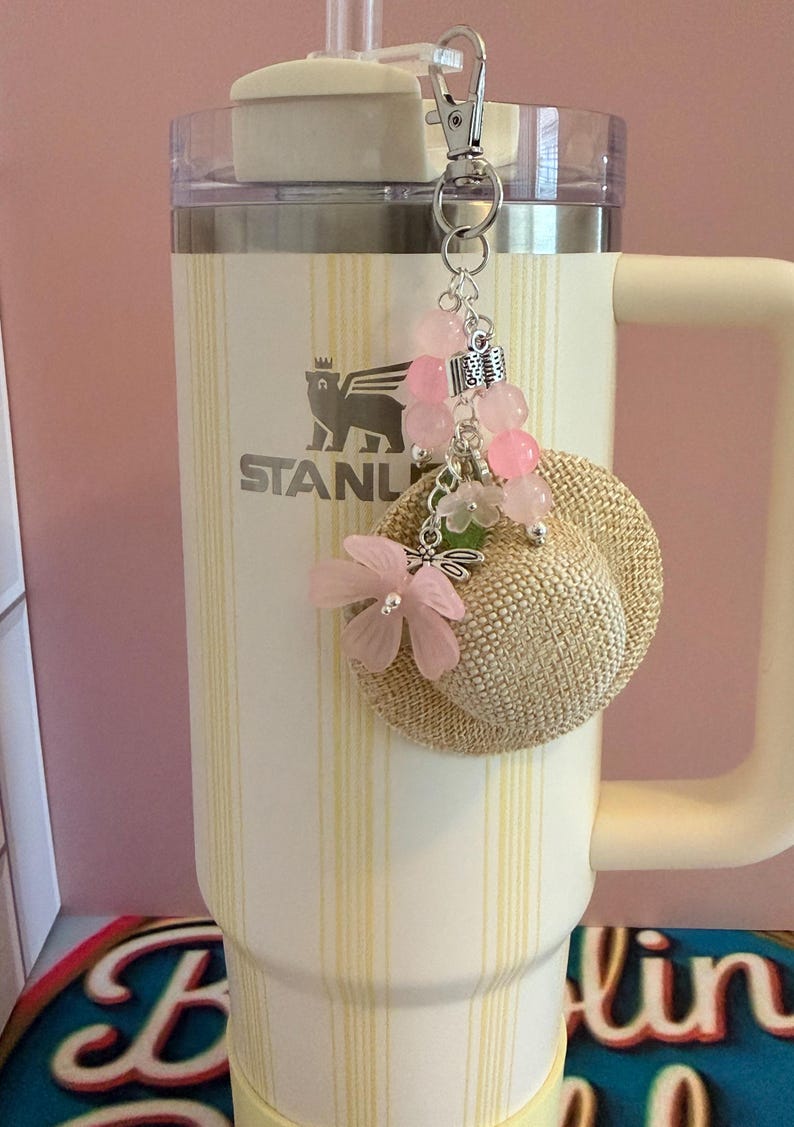 May include: A cream-coloured Stanley tumbler with a straw and a decorative keychain. The keychain features a small straw hat, pink flower accents, and pink bead embellishments. The tumbler has the Stanley logo and a yellow striped pattern.