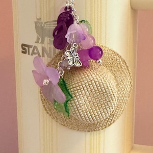 May include: A decorative keychain with a small straw hat decorated with purple flowers, beads, and a silver butterfly charm. The keychain also includes purple flip-flops and is attached to a silver ring.