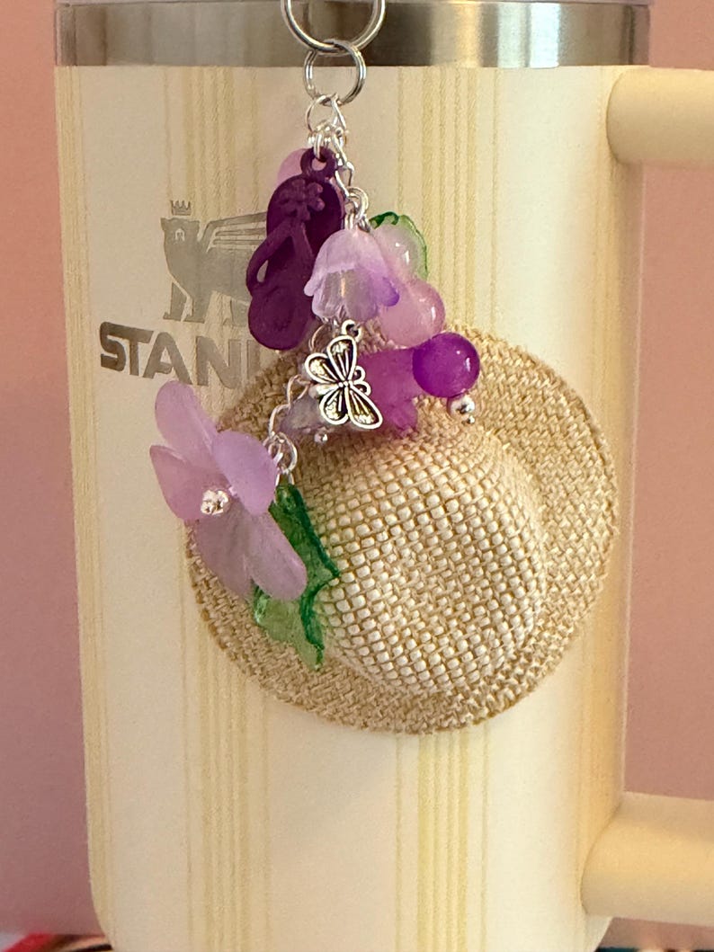May include: A decorative keychain with a small straw hat decorated with purple flowers, beads, and a silver butterfly charm. The keychain also includes purple flip-flops and is attached to a silver ring.
