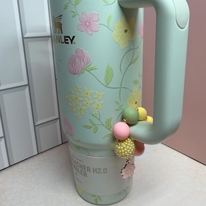May include: A mint green Stanley Quencher H2.0 tumbler with a floral design featuring pink and yellow flowers. The tumbler has a handle and a decorative charm with pink, yellow, and green beads. The Stanley logo is visible.