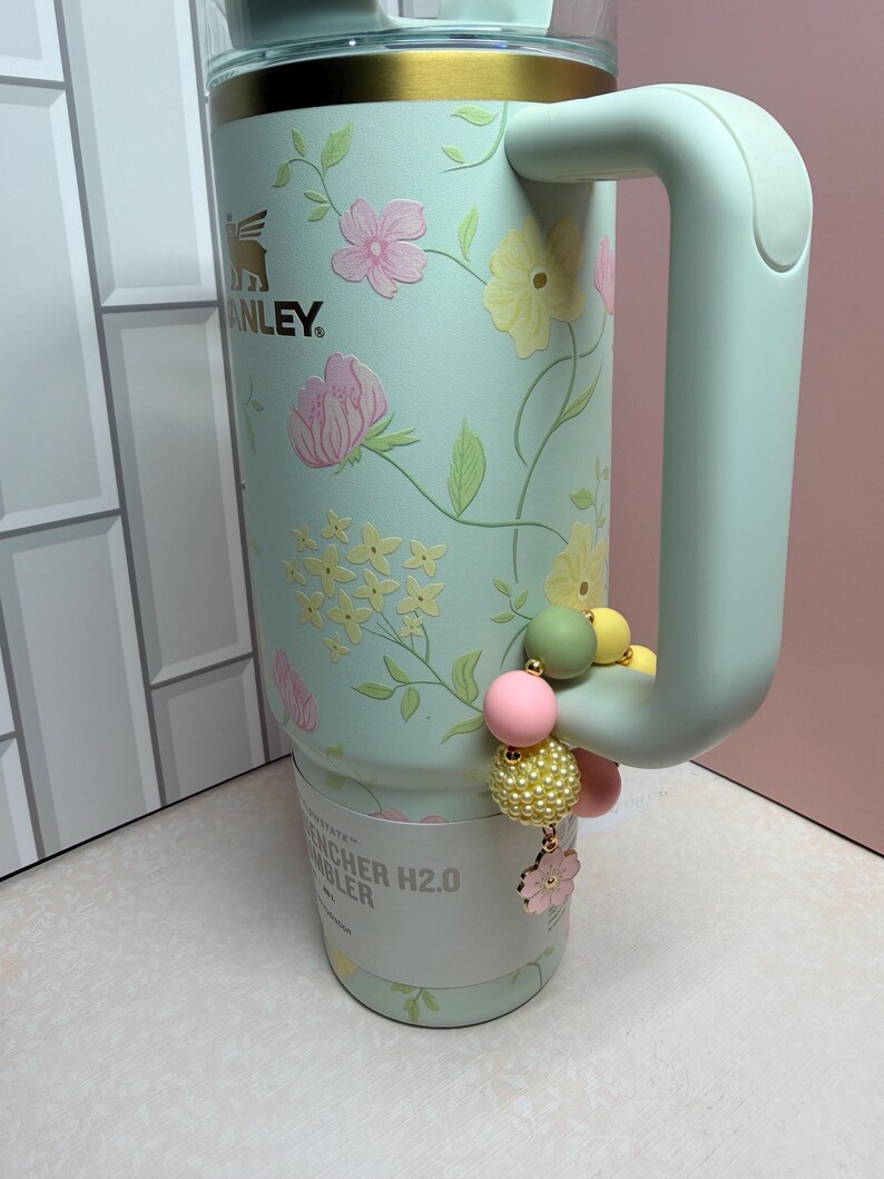 May include: A mint green Stanley Quencher H2.0 tumbler with a floral design featuring pink and yellow flowers. The tumbler has a handle and a decorative charm with pink, yellow, and green beads. The Stanley logo is visible.