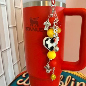 Puede incluir: Un vaso t&eacute;rmico de color rojo brillante con asa y una base a cuadros blancos y negros. El logotipo de Stanley est&aacute; impreso en el frente. Un dije decorativo cuelga de la tapa, con cuentas de tem&aacute;tica de f&uacute;tbol y las letras ESP.