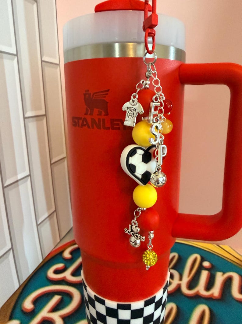 Puede incluir: Un vaso t&eacute;rmico de color rojo brillante con asa y una base a cuadros blancos y negros. El logotipo de Stanley est&aacute; impreso en el frente. Un dije decorativo cuelga de la tapa, con cuentas de tem&aacute;tica de f&uacute;tbol y las letras ESP.