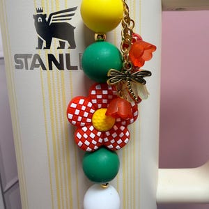 May include: A decorative charm featuring a yellow bead, a green bead, a red and white checkered flower bead, another green bead, and a white bead. It includes a gold-toned dragonfly charm and small orange flower accents on a gold chain.