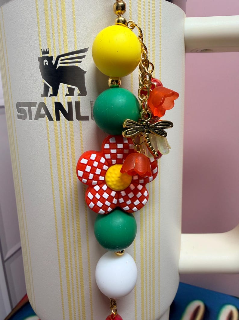 May include: A decorative charm featuring a yellow bead, a green bead, a red and white checkered flower bead, another green bead, and a white bead. It includes a gold-toned dragonfly charm and small orange flower accents on a gold chain.