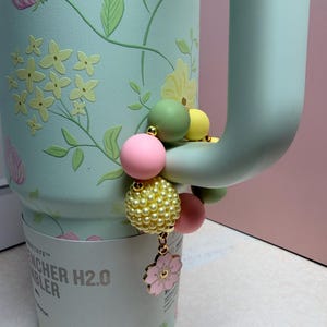 May include: A mint green tumbler with a floral design and a beaded charm. The charm features pink, yellow, and green beads, with a pink flower pendant. The tumbler has a handle and the text "STANLEY" and "H2.0" is visible.