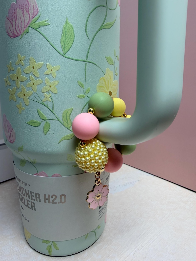 May include: A mint green tumbler with a floral design and a beaded charm. The charm features pink, yellow, and green beads, with a pink flower pendant. The tumbler has a handle and the text "STANLEY" and "H2.0" is visible.