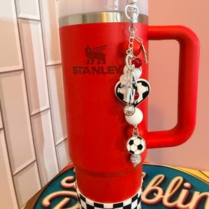 Puede incluir: Un vaso Stanley de color rojo brillante con asa y tapa blanca, que presenta una base a cuadros blancos y negros. Un dije decorativo cuelga de la tapa, mostrando cuentas rojas y blancas, un dije de bal&oacute;n de f&uacute;tbol y un colgante de coraz&oacute;n.