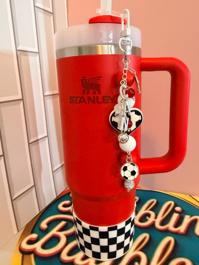 Puede incluir: Un vaso Stanley de color rojo brillante con asa y tapa blanca, que presenta una base a cuadros blancos y negros. Un dije decorativo cuelga de la tapa, mostrando cuentas rojas y blancas, un dije de bal&oacute;n de f&uacute;tbol y un colgante de coraz&oacute;n.