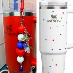 Op de afbeelding: Rode Stanley-beker met een bedel met rode, witte en blauwe kralen en een bloemaccent. De beker heeft het Stanley-logo. Een witte Stanley-mok met een handvat is versierd met rode, blauwe en grijze sterren.