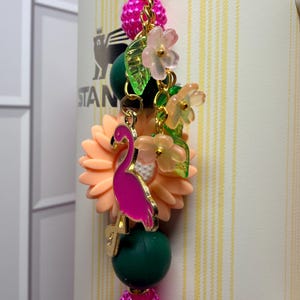 May include: A decorative tumbler charm featuring a bright pink flamingo pendant, a large peach-colored sunflower bead, dark green round beads, and textured hot pink beads. The charm includes small translucent flower and leaf accents on gold links.