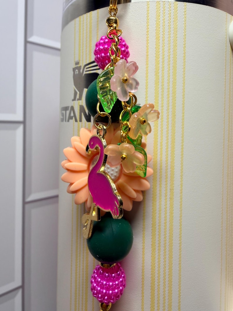 May include: A decorative tumbler charm featuring a bright pink flamingo pendant, a large peach-colored sunflower bead, dark green round beads, and textured hot pink beads. The charm includes small translucent flower and leaf accents on gold links.