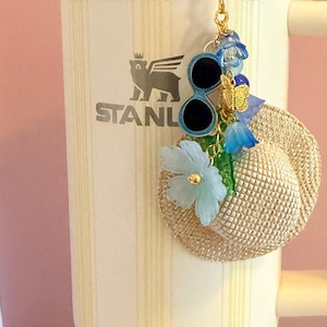 May include: A cream Stanley tumbler with a gold keychain. The keychain features a miniature straw hat, sunglasses, a butterfly, and blue floral accents. The tumbler has a vertical striped pattern.