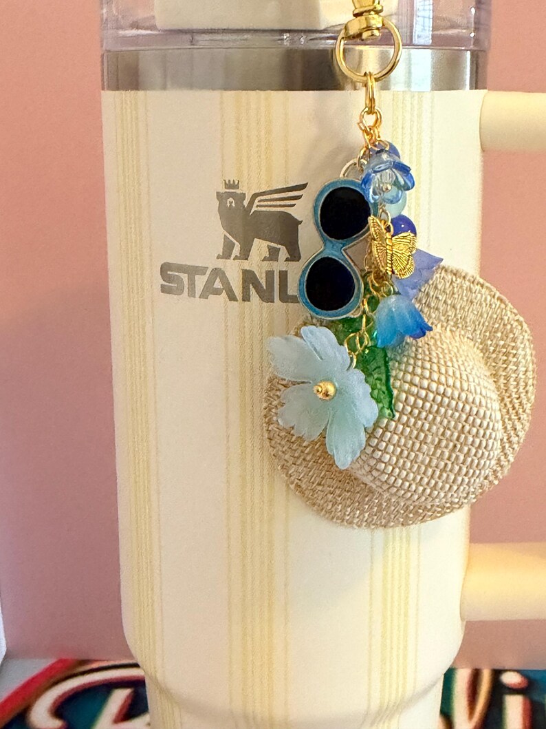 May include: A cream Stanley tumbler with a gold keychain. The keychain features a miniature straw hat, sunglasses, a butterfly, and blue floral accents. The tumbler has a vertical striped pattern.