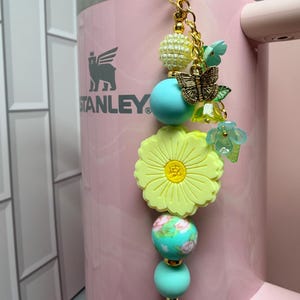 May include: A decorative tumbler charm featuring a gold-tone metal butterfly, a large yellow flower bead, and various round beads in shades of turquoise, cream, and floral patterns. The charm hangs from a gold-tone chain attached to a light pink tumbler.