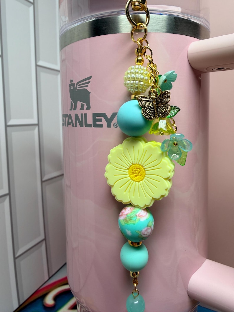May include: A decorative tumbler charm featuring a gold-tone metal butterfly, a large yellow flower bead, and various round beads in shades of turquoise, cream, and floral patterns. The charm hangs from a gold-tone chain attached to a light pink tumbler.