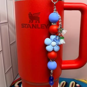 Op de afbeelding: Een rode Stanley-beker met een handvat en een blauwe rietje. Een decoratieve bedel met blauwe en rode kralen, bloemen en een zilveren sluiting hangt aan het handvat van de beker. Het Stanley-logo is zichtbaar.
