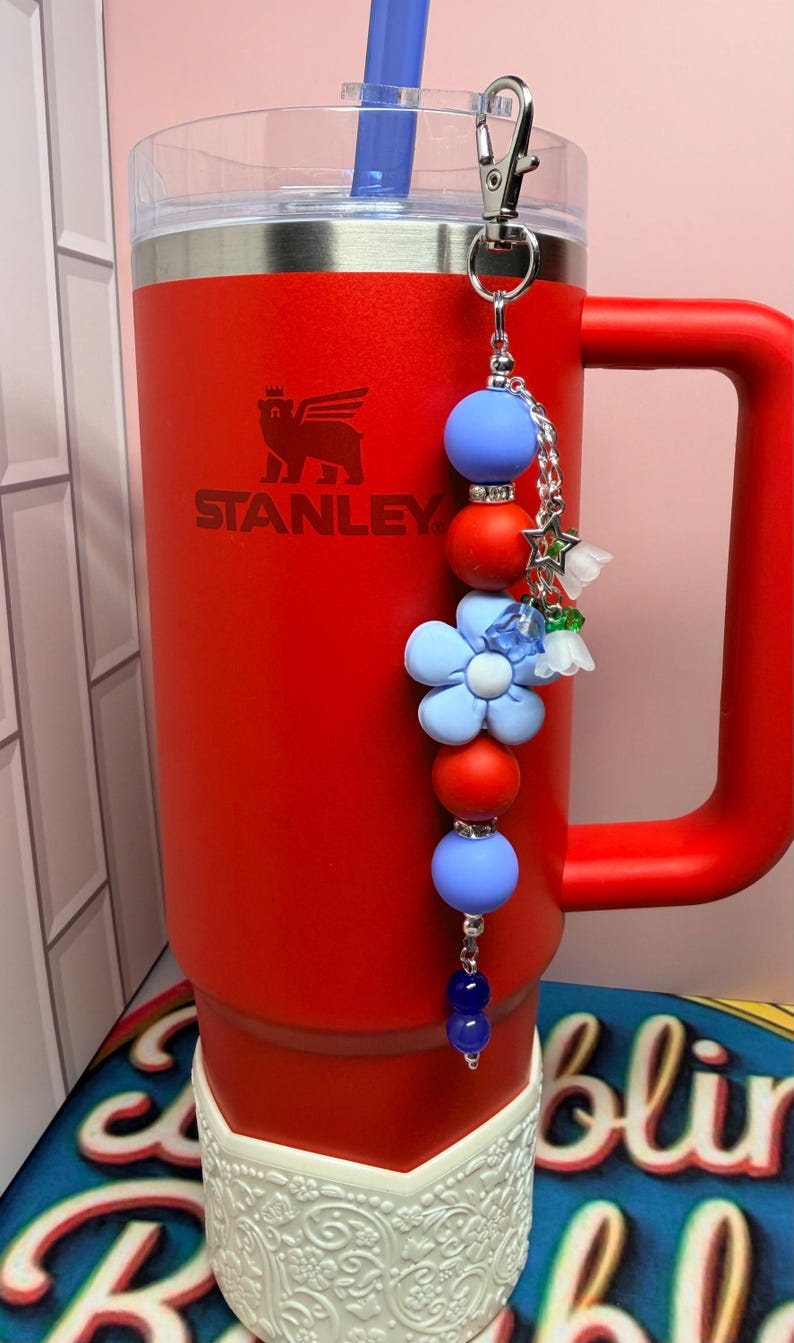 Op de afbeelding: Een rode Stanley-beker met een handvat en een blauwe rietje. Een decoratieve bedel met blauwe en rode kralen, bloemen en een zilveren sluiting hangt aan het handvat van de beker. Het Stanley-logo is zichtbaar.
