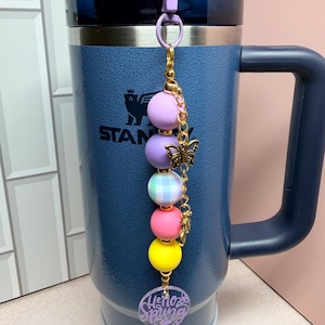 May include: A blue insulated travel mug with a handle and a decorative charm. The charm features a series of colourful beads, a gold butterfly, and a charm that says "Hello Spring". The mug has the word "STANLEY" printed on it.