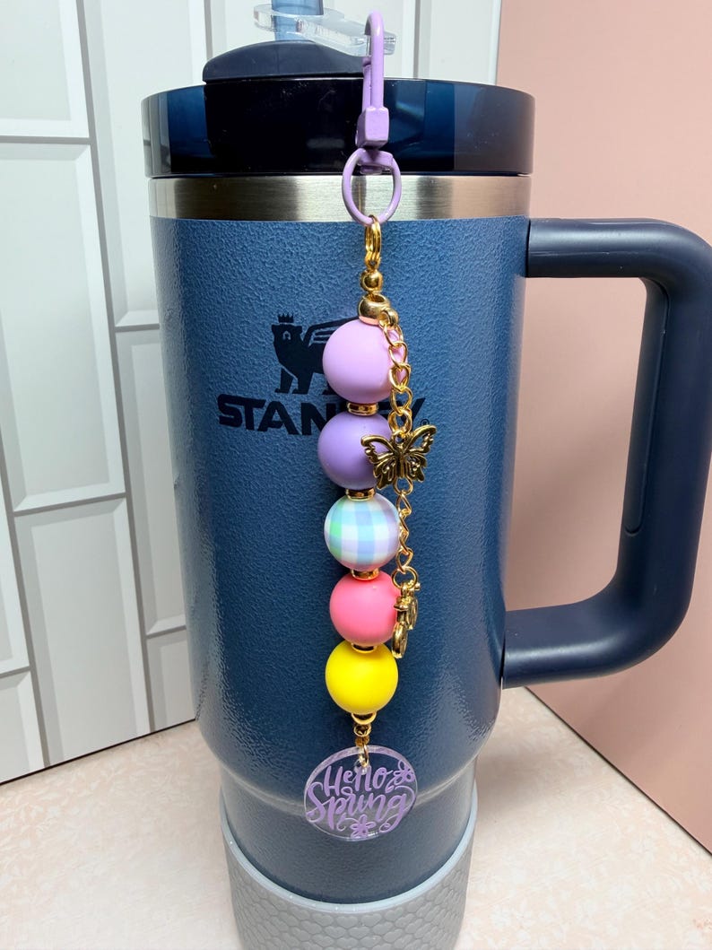 May include: A blue insulated travel mug with a handle and a decorative charm. The charm features a series of colourful beads, a gold butterfly, and a charm that says "Hello Spring". The mug has the word "STANLEY" printed on it.