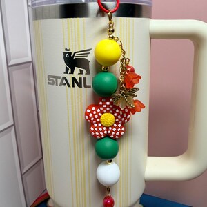 May include: A cream-colored Stanley tumbler with a vertical yellow striped pattern features a decorative beaded charm attached to the handle. The charm includes yellow, green, and white beads, a red and white checkered flower, and a gold dragonfly charm.