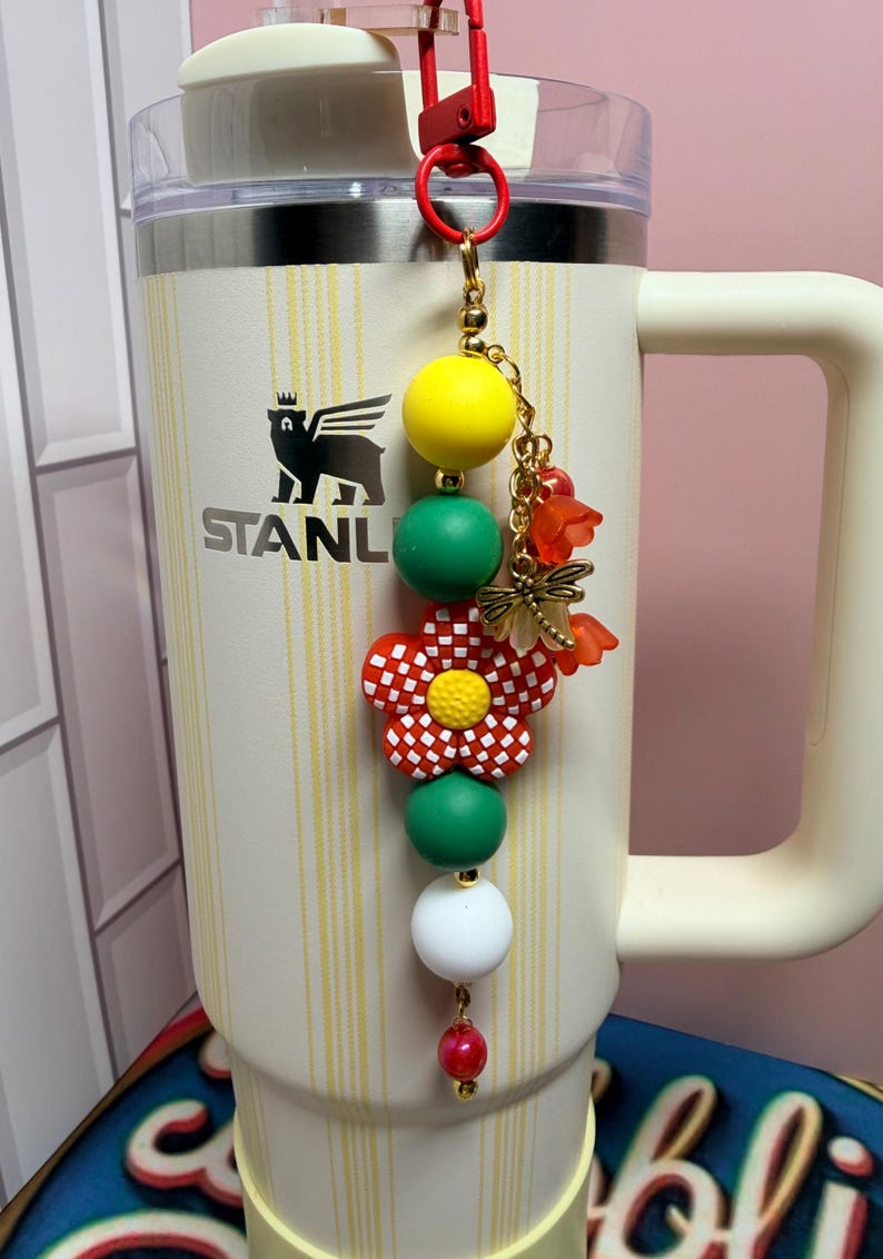 May include: A cream-colored Stanley tumbler with a vertical yellow striped pattern features a decorative beaded charm attached to the handle. The charm includes yellow, green, and white beads, a red and white checkered flower, and a gold dragonfly charm.