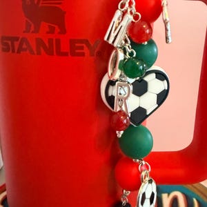 Puede incluir: Un vaso Stanley rojo con un accesorio decorativo. El dije consiste en una cadena plateada con cuentas rojas, verdes y negras, letras plateadas que forman la palabra MOR, un dije de coraz&oacute;n con dise&ntilde;o de bal&oacute;n de f&uacute;tbol y un peque&ntilde;o colgante de bal&oacute;n.