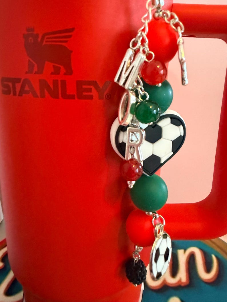 Puede incluir: Un vaso Stanley rojo con un accesorio decorativo. El dije consiste en una cadena plateada con cuentas rojas, verdes y negras, letras plateadas que forman la palabra MOR, un dije de coraz&oacute;n con dise&ntilde;o de bal&oacute;n de f&uacute;tbol y un peque&ntilde;o colgante de bal&oacute;n.